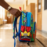 A colorful children's backpack with a cartoon dinosaur pattern and a red front pocket featuring a yellow 'CARTER' emergency contact patch, being held by a person's hand in a school hallway.