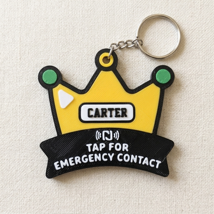 A novelty crown-shaped keychain with a yellow crown body, black outline, green circular accents, a white nameplate reading 'CARTER,' and a black banner below that says 'TAP FOR EMERGENCY CONTACT.' The keychain is photographed on a light beige fabric background.